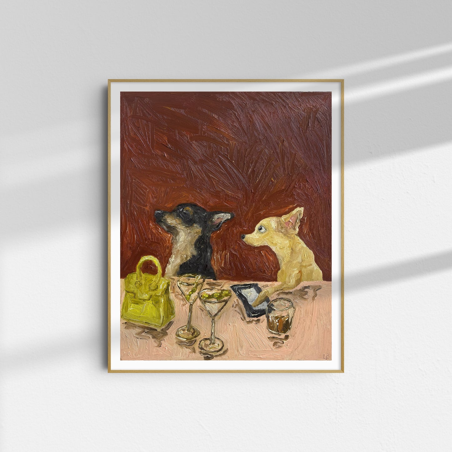 A framed oil on canvas reproduction of Leah Boyd's "It's Not on Me" depicts two small dogs seated at a table with two martini glasses, a yellow handbag, and a phone against a textured red background. Sunlight casts shadows on the wall behind the artwork.