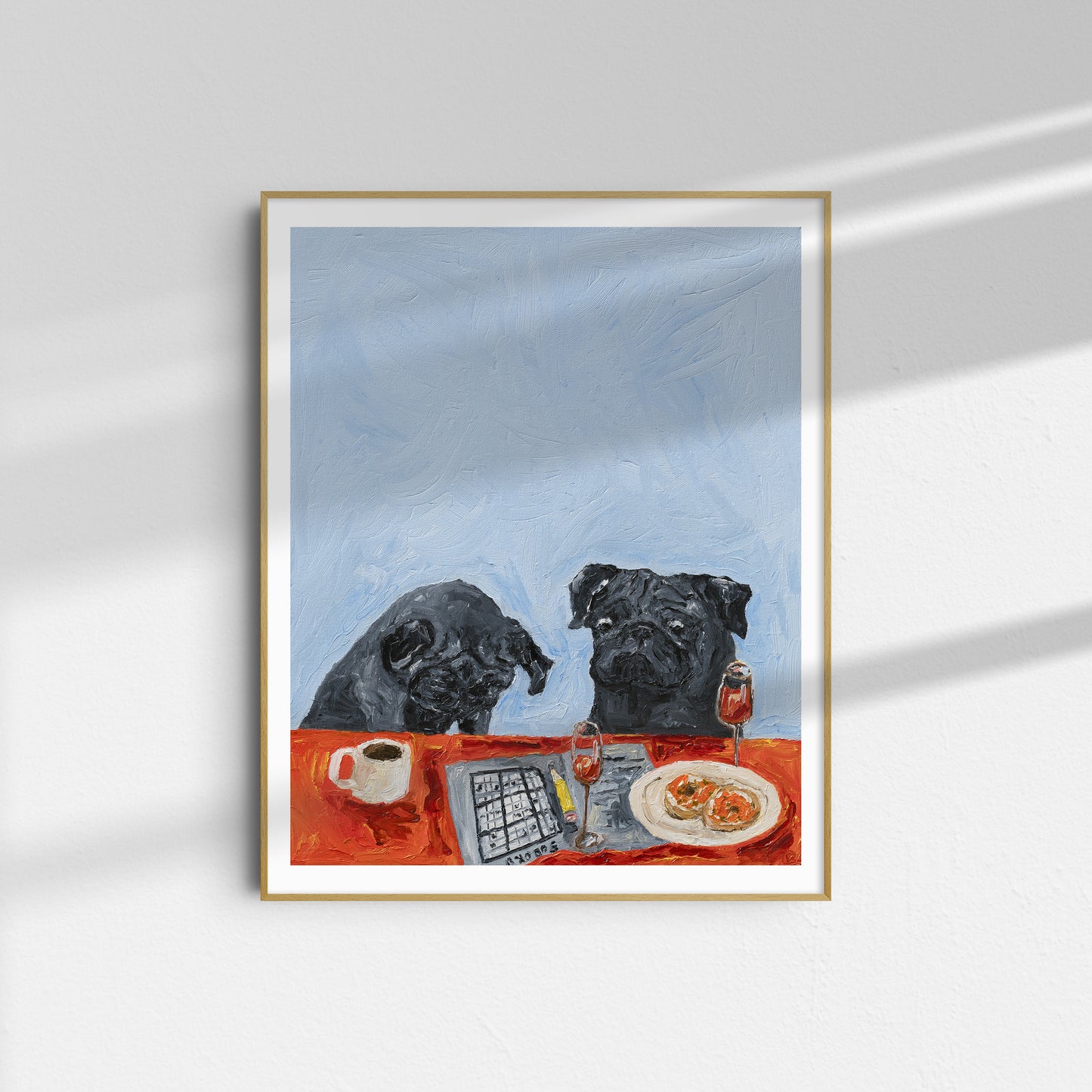 A framed limited edition Giclee print titled “Level Hard Sudoku” by Leah Boyd, featuring two black dogs sitting at a table with food, a coffee cup, and a wine glass against a light blue background, hangs on a white wall with sunlight casting shadows.
