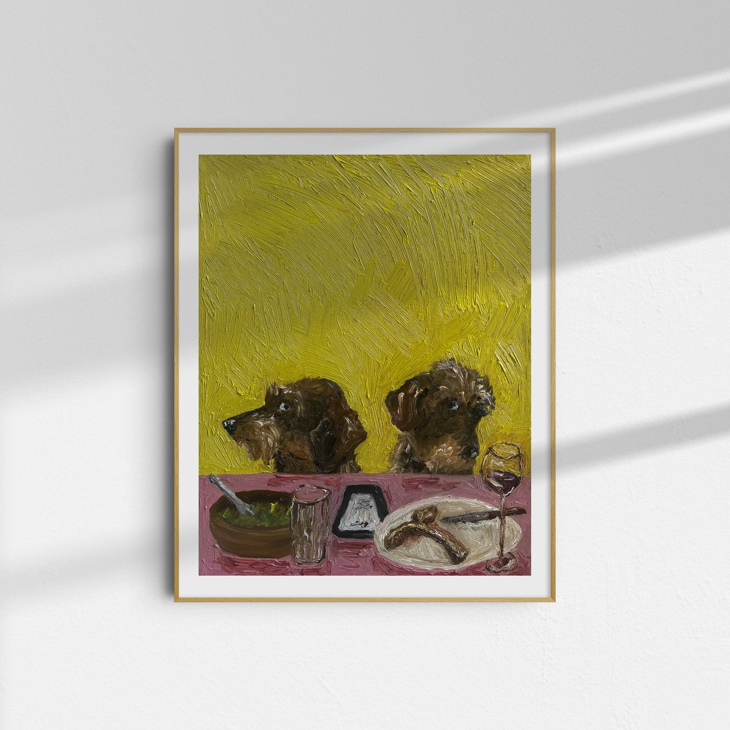 Limited edition "We're Not Splitting" by Leah Boyd—this framed giclée print features two brown dogs seated at a pink table set with dishes, a wine glass, and food, against a textured yellow background. Displayed on a white wall with soft shadows.