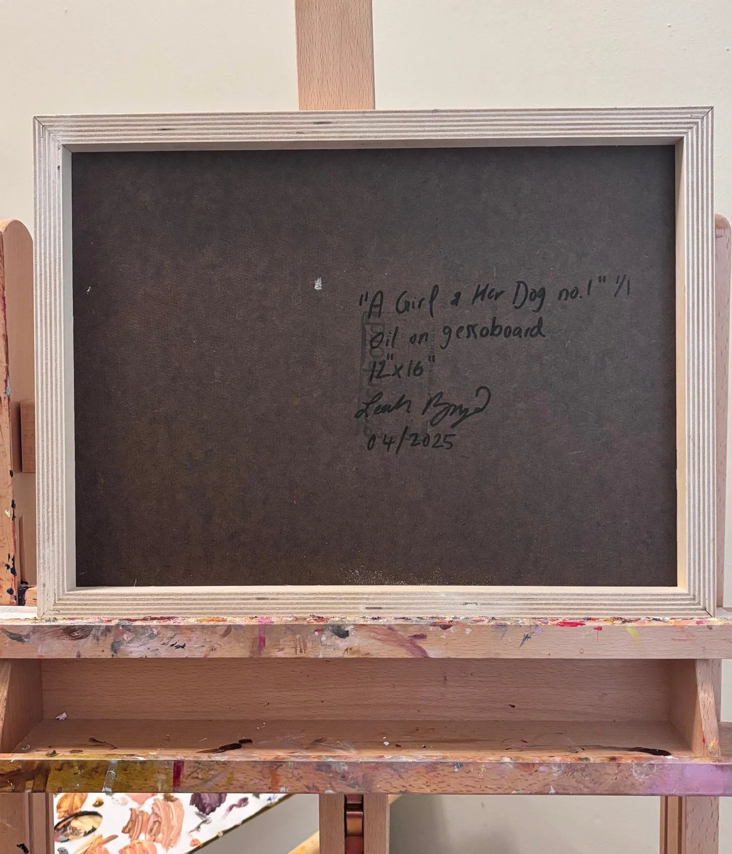 A framed, blank dark board rests on a wooden easel. The back features handwritten text detailing "A Girl and Her Dog" by Leah Boyd, an original signed 12x16 oil painting on gessoboard, dated 04/2025.