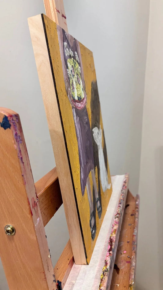 Leah Boyd’s "A Girl & Her Dog no. 2" is a cradled gesso-board oil painting on a wooden easel, depicting a side view of a figure with flowers, set against a plain light wall.