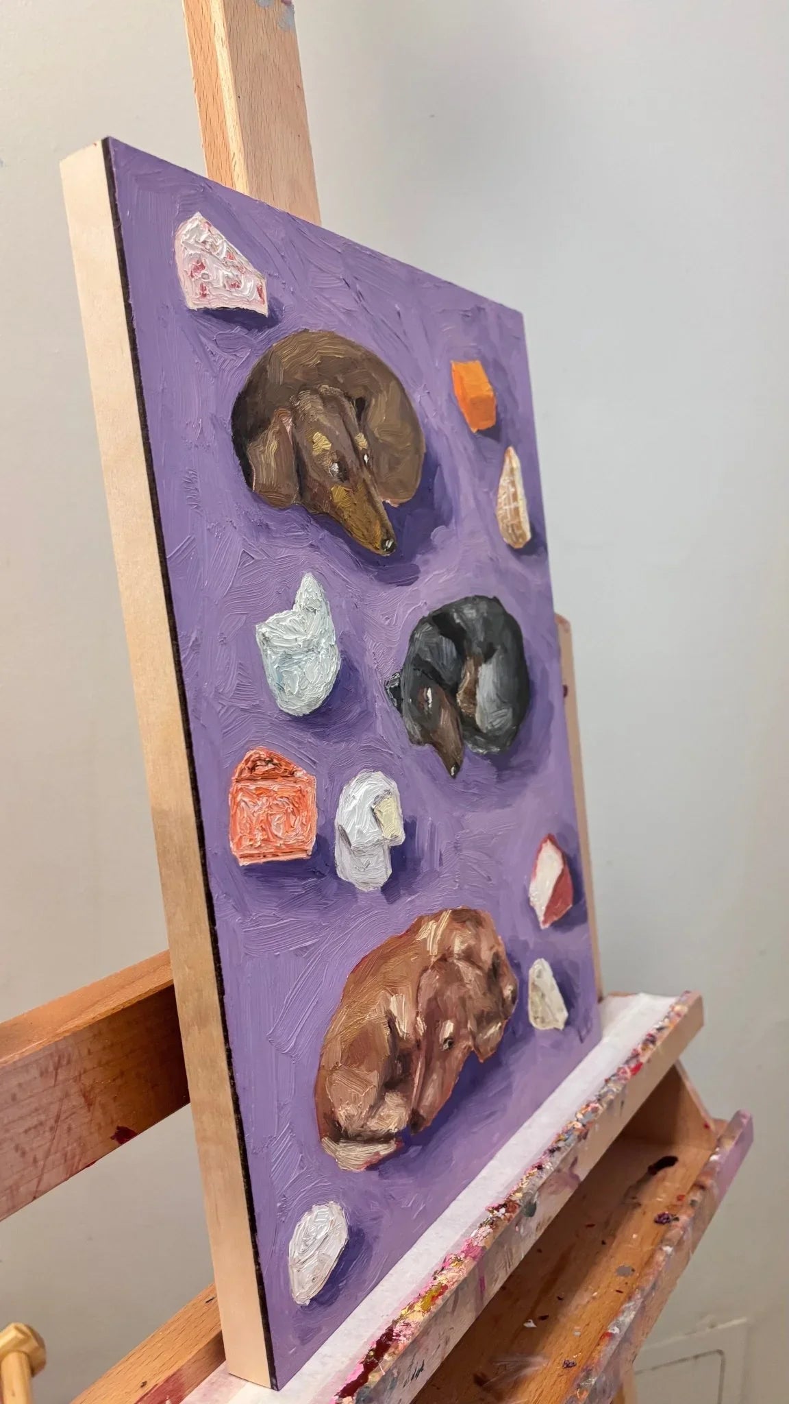 "Dachshunds and Cheese" by Leah Boyd is an original oil painting on gesso-board, featuring three sleeping dachshunds and cheese wedges on a purple background, with thick texture visible from the side.