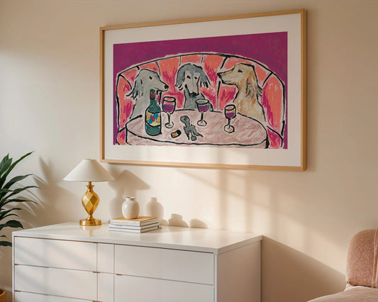 A framed Giclee print, "Wine Night & Yap" by Leah Boyd, depicting three dogs with wine glasses and a bottle, hangs above a white dresser with a gold lamp, two books, and vases in a bright room.