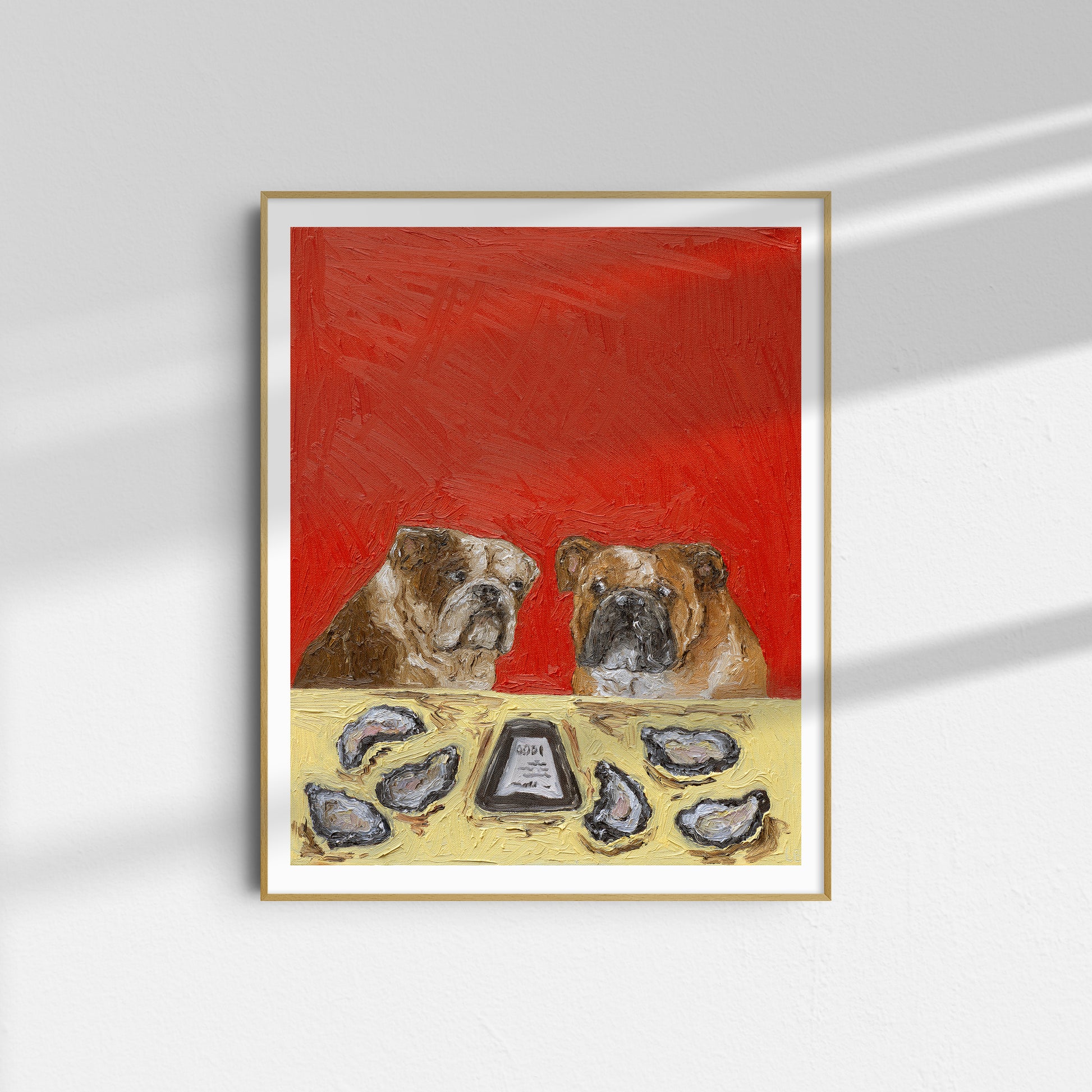 A framed limited edition art print titled “$1 Oysters Ended at 6” by Leah Boyd features two bulldogs seated at a table with oyster shells and a bottle labeled cocktail sauce on a red background, displayed on a white wall with soft light and shadow patterns.