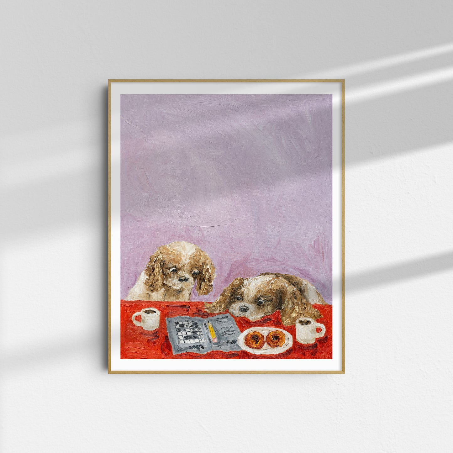 A framed limited edition art print titled “Cavalier Conundrum” by Leah Boyd, depicting two dogs sitting at a table with coffee cups, a newspaper, and pastries against a light purple background, hangs on a white wall.