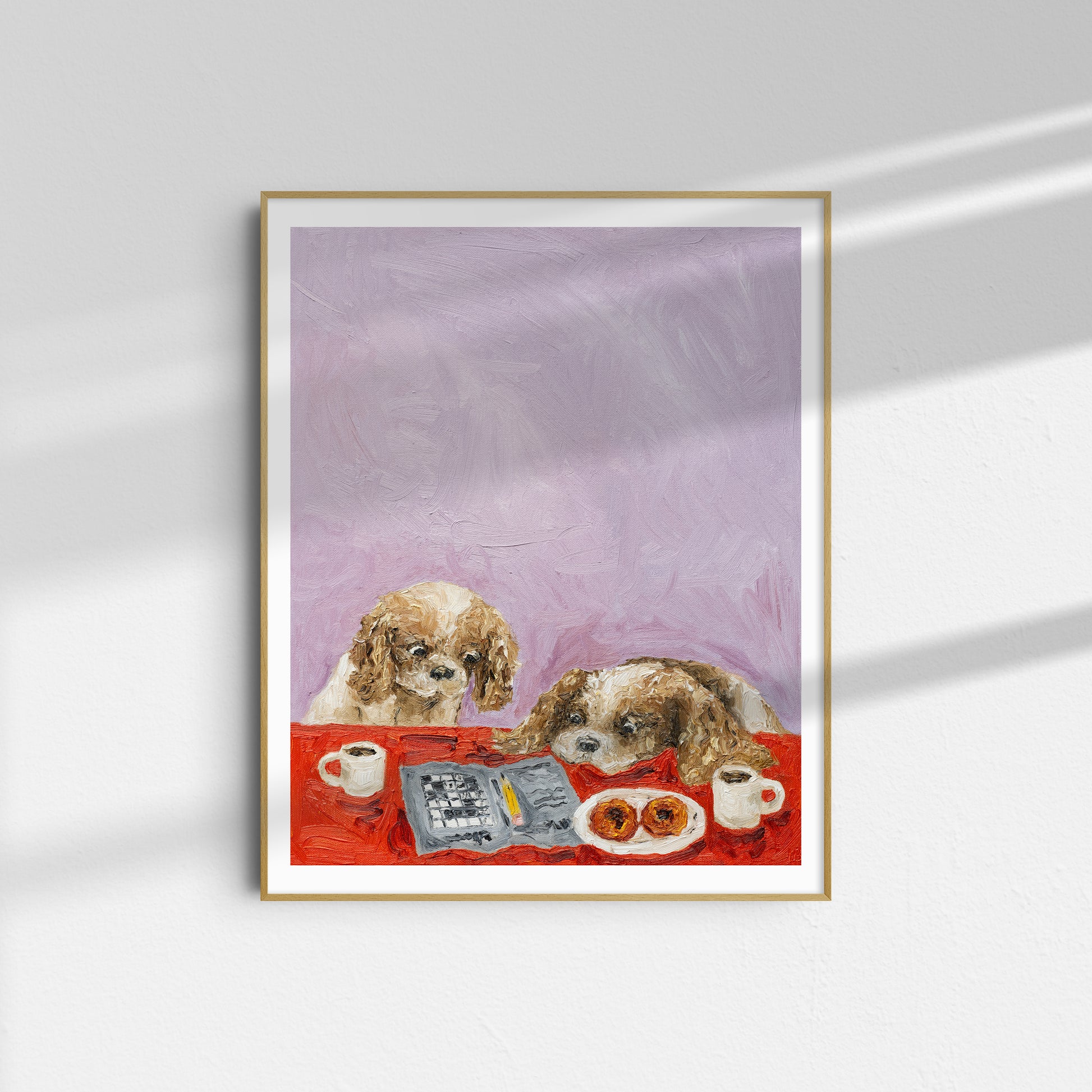 A framed limited edition art print titled “Cavalier Conundrum” by Leah Boyd, depicting two dogs sitting at a table with coffee cups, a newspaper, and pastries against a light purple background, hangs on a white wall.