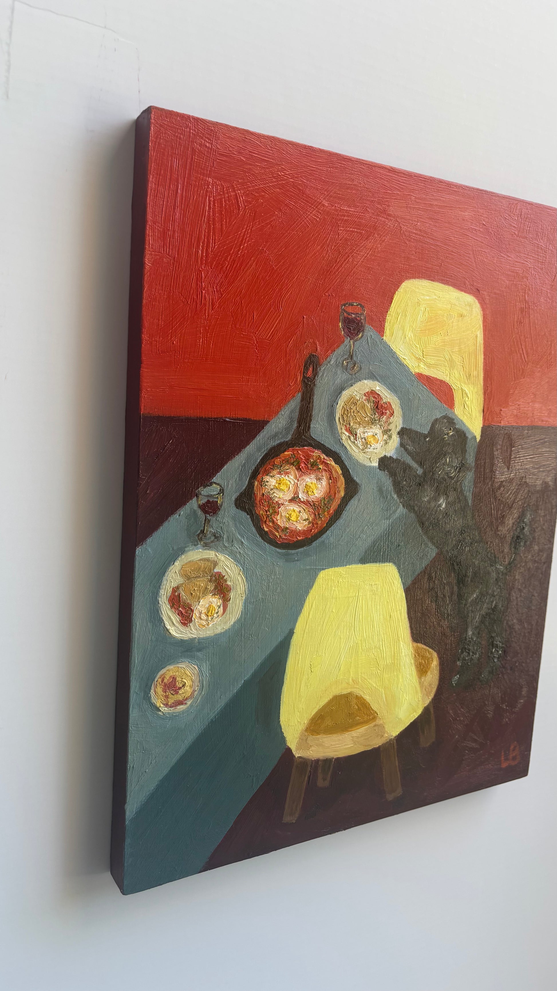 Dinner for One no. 5: Shakshuka by Leah Boyd is a vibrant oil painting on cradled wood panel featuring bold reds, blues, and yellows, a set table with food and wine, two yellow chairs, and a black cat reaching for a plate.