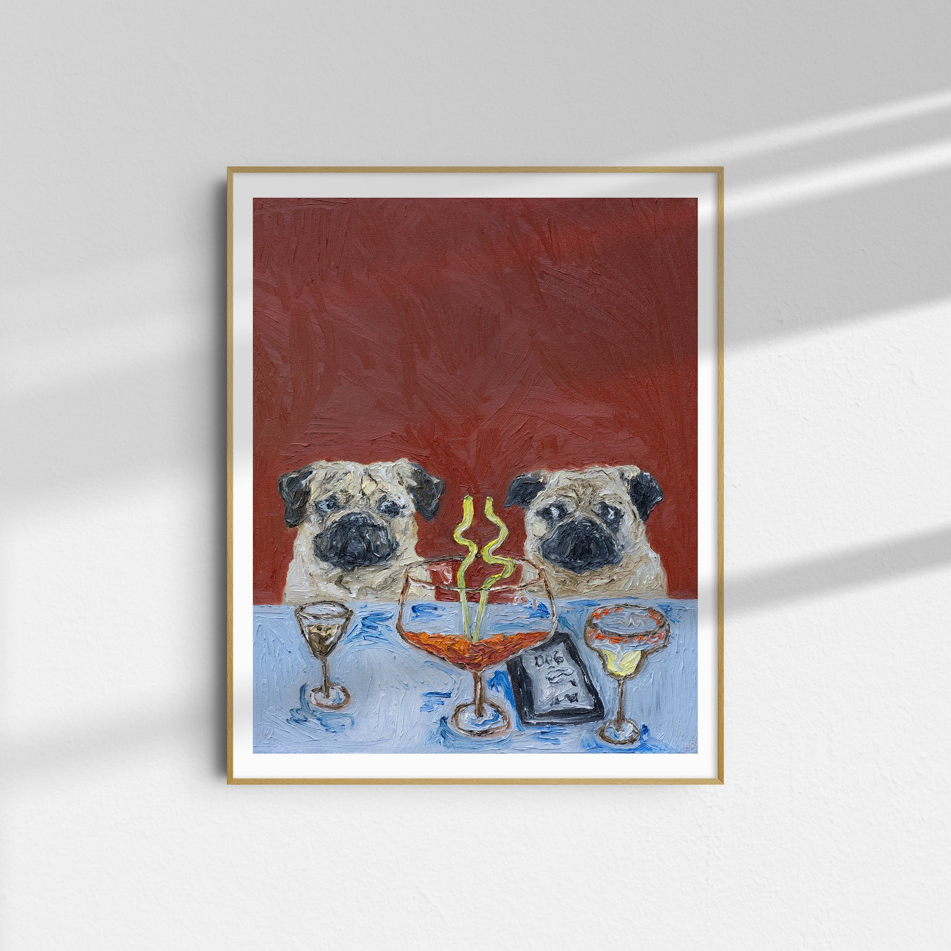 Framed oil on canvas painting titled “Who’s Driving?” by Leah Boyd, featuring two pugs sitting at a table with drinks, a dessert, and a menu that reads DOG, set against a textured red background. The artwork hangs on a white wall with sunlight casting shadows.